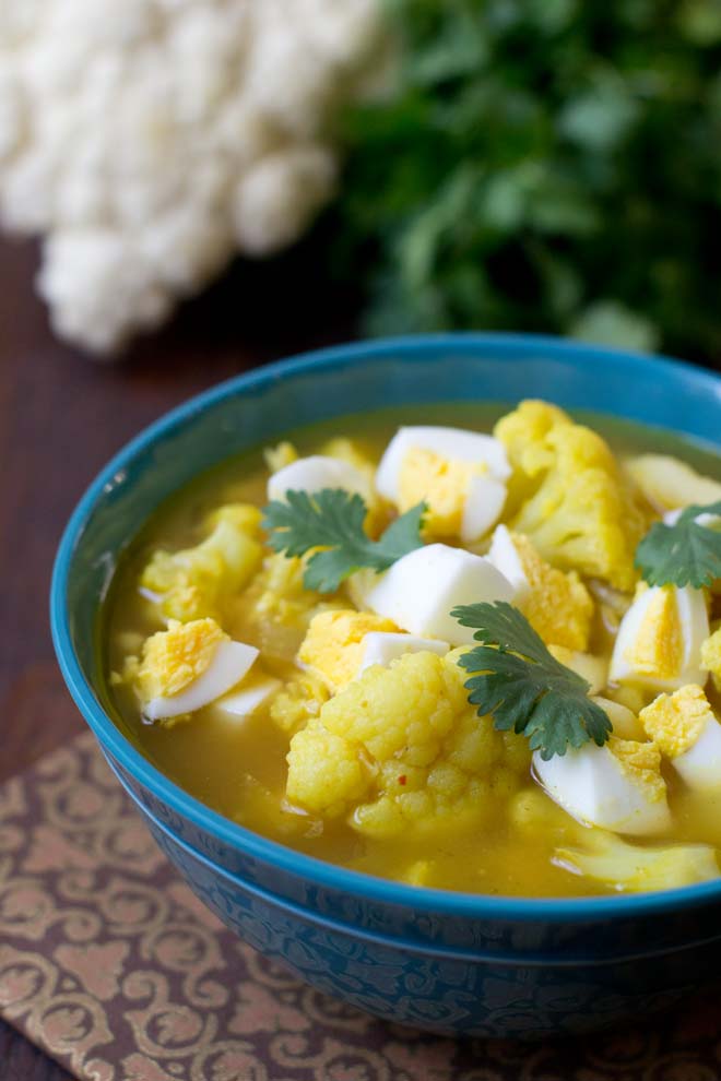 Indian-Spiced Cauliflower Soup with chopped hard boiled eggs garnished with cilantro leaves in a blue bowl; in the background there is a bunch of cilantro and head of cauliflower.