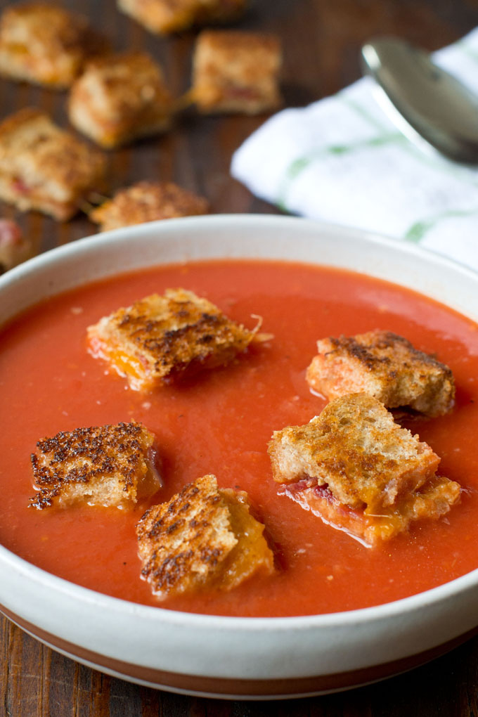 Tomato Soup with Grilled Cheese and Bacon Croutons in a white soup bowl; in the background is a white and green checked cloth napkin with a spoon sitting on it and next to the napkin is some cheese and bacon croutons.