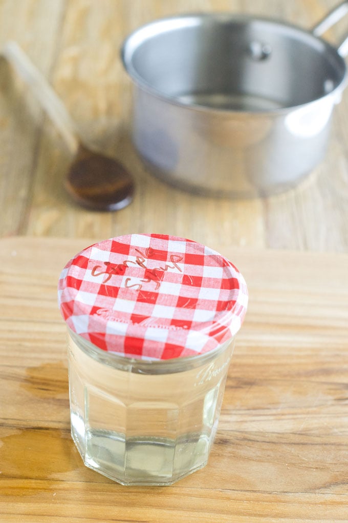 Clear jar with red and white checkered top with the words, "Simple Syrup" written on it, filled with clear simple syrup. In the background is a silver pot and wooden spoon.