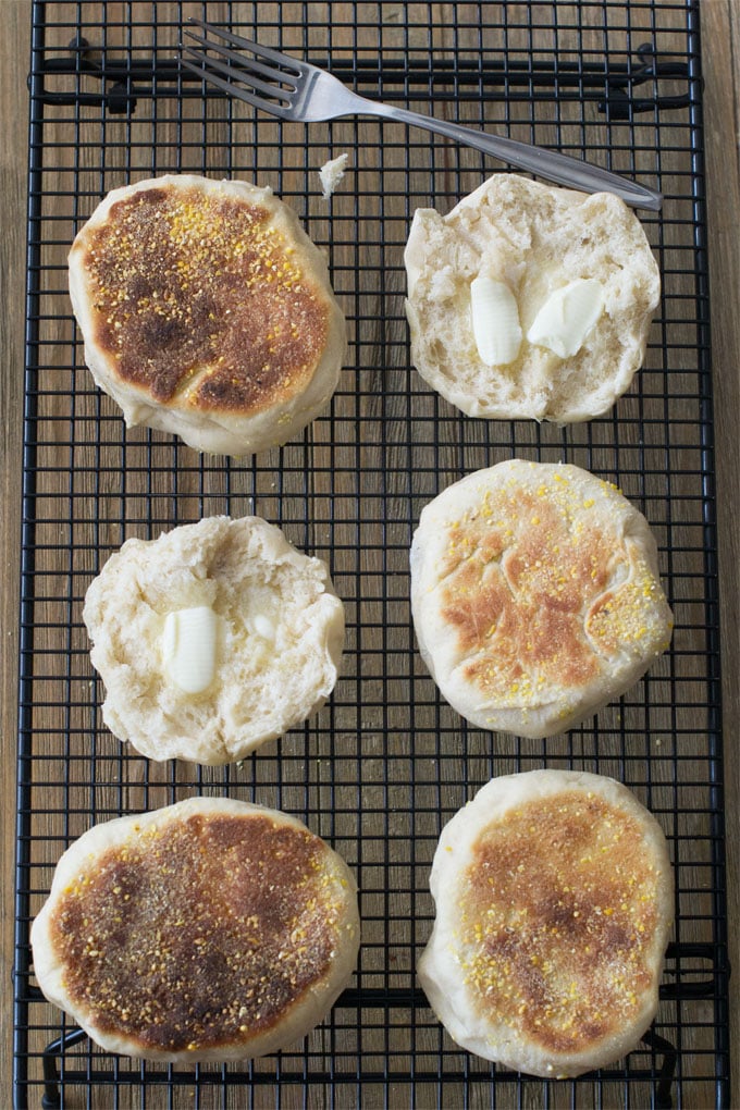 Toasted English muffin halves on a black wire cooling rack. Some are turned upside down. The halves that are turned right side up have half melted pats of butter on them. There is a fork on the cooling rack as well.