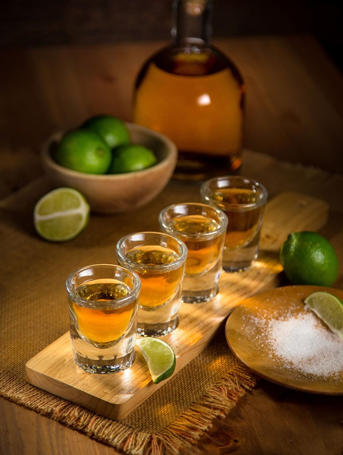 4 shot glasses on a wooden board filled with tequila. There is a plate of salt with a lime wedge on an and a full lime to the right of the board. In the background is a half of a lime, a wooden bowl full of whole limes and a glass bottle full of light brown tequila.