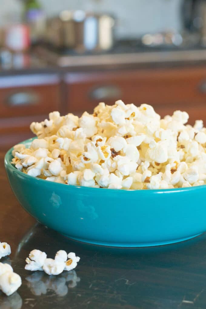 A blue bowl full to overflowing with popcorn. Some kernels are on the counter beside it. There's a stove with a metal pot on it in the background.