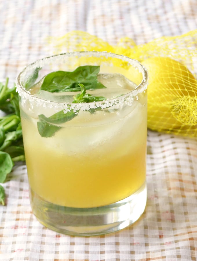 Short glass with salted rim and filled with lemon margarita, ice and topped with whole basil leaves. It is sitting on a white checkered tablecloth with some basil leaves and a mesh bag of lemons behind the glass.