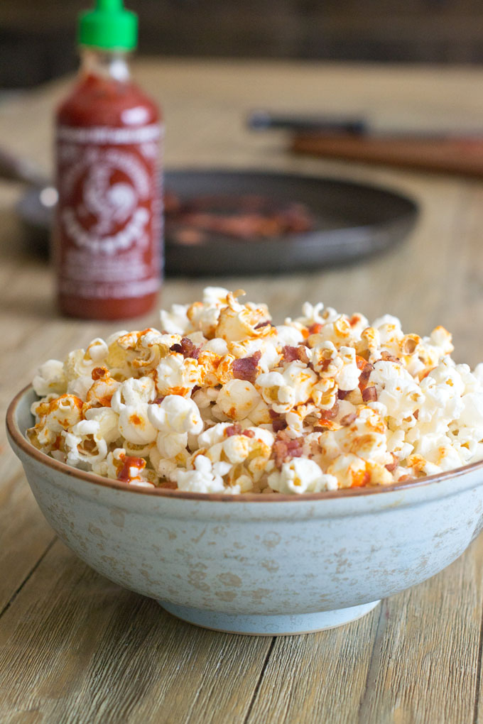 Sriracha Bacon Popcorn in a bowl on a wood table with a bottle of Sriracha and plate of bacon in the background.