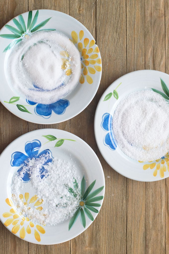 Three plates with blue, yellow and green flowers with a different kind of salt on each one.