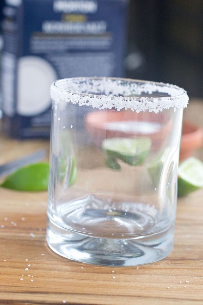 Short clear class with salted rim sitting on a wood surface with wedges of lime and a box of salt behind it.