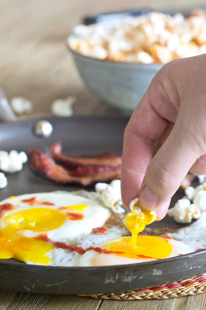 Soft fried eggs drizzled with hot sauce and strips of cooked bacon in a frying pan with scattered pieces of popcorn; a hand is dipping a piece of popcorn in the runny yolk.