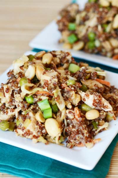 Thai Peanut Salad with Quinoa, peanuts, green onions , chicken and cabbage piled high on a small square plate with an identical plate in the background. The plates are sitting on a blue cloth napkin on a wooden table.