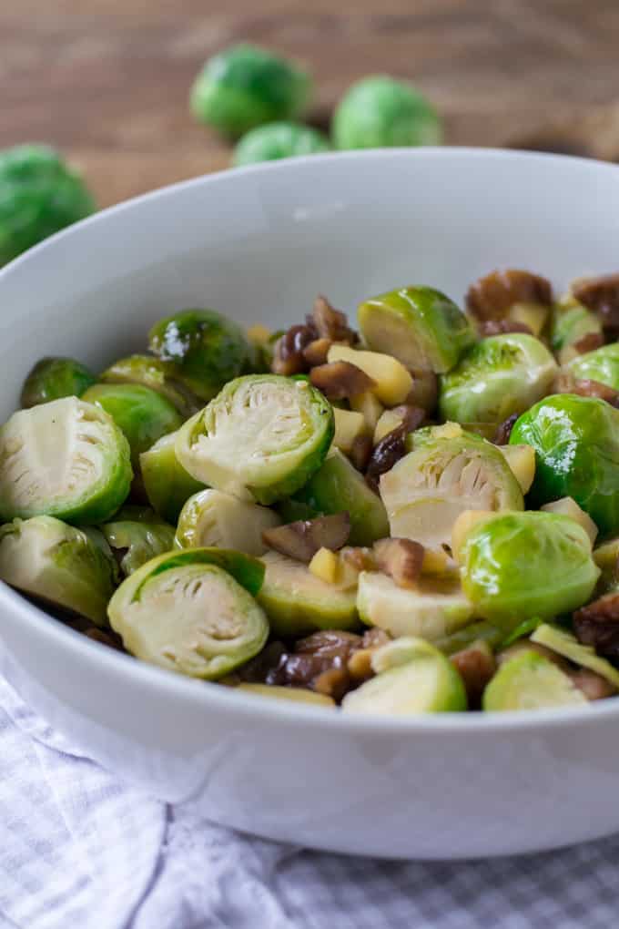White bowl filled with halved Brussels sprouts that have been sautéed with apples and chestnuts. The bowls is sitting on a grey checked dish towel on a wood table with some whole Brussels sprouts in the background.