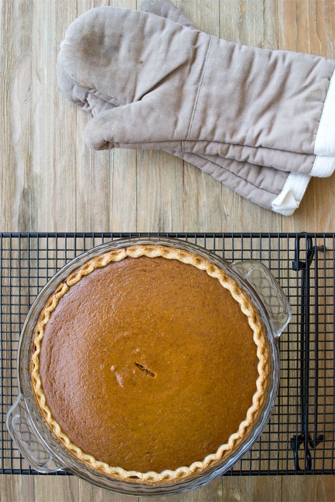 Classic Pumpkin Pie in a glass pie dish on a cooling rack, pair of oven mitts beside.