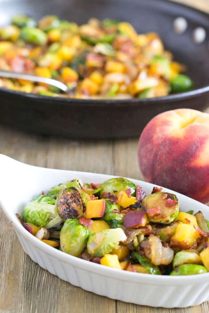 White oblong gratin dish on a wooden table filled with Brussels sprouts cooked with peaches and bacon. A whole peach is sitting behind the dish and, blurred in the background, is a black skillet filled with Brussels sprouts cooked with peaches and bacon.