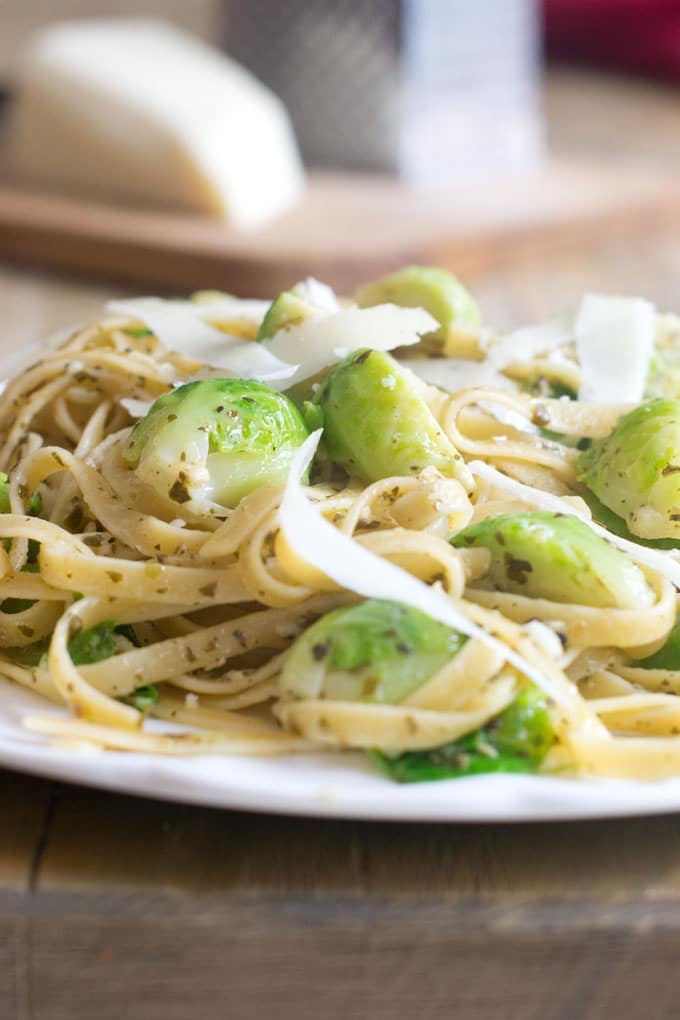 Close up of fettuccni pasta with Brussels sprouts and shaved pecorino cheese on a white plate on a  wood table with a cheese grater and wedge of white cheese in the background on a wood cutting board.
