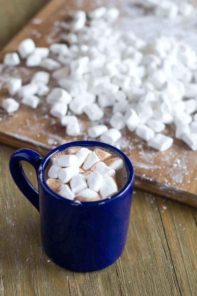 Blue mug of hot chocolate with marshmallows, cutting board filled with fresh marshmallows in background.