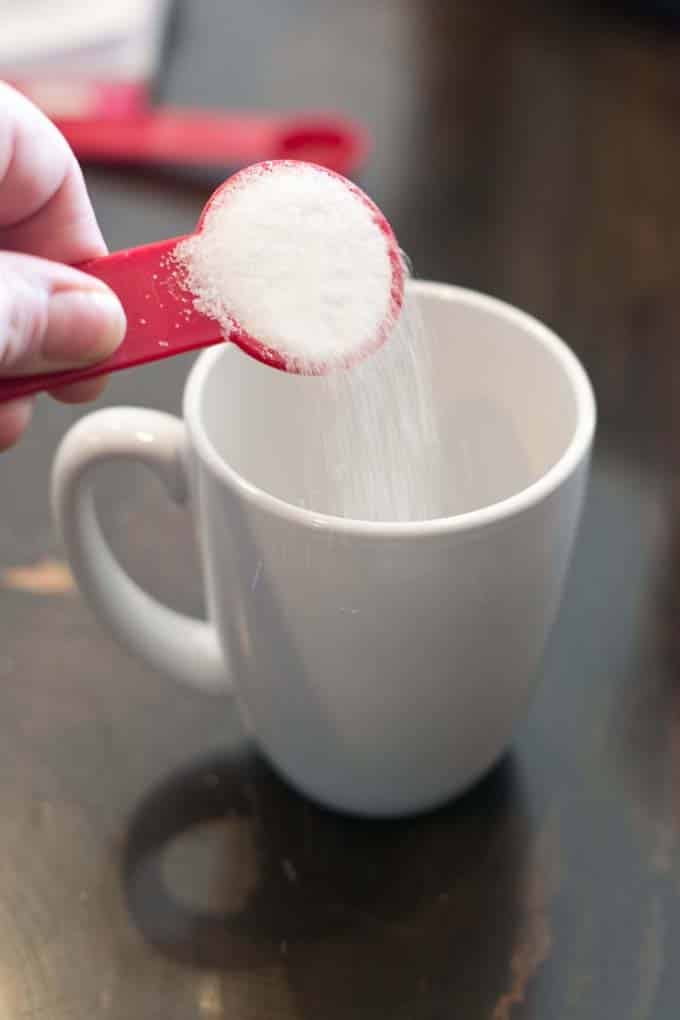 Red measuring spoon of sugar being poured into white mug.