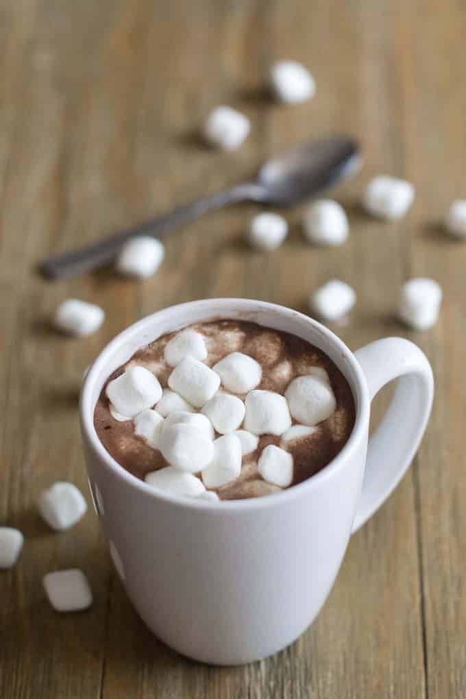 White mug filled with hot cocoa and mini marshmallows. The table it is sitting has mini marshmallows scattered on it and a spoon.