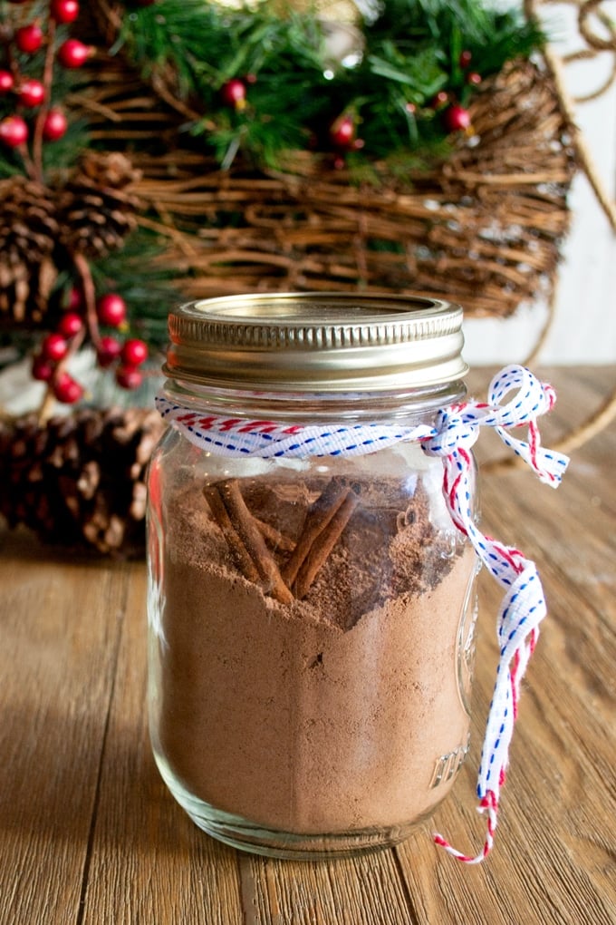 Mason Jar filled with hot chocolate mix and two cinnamon sticks. There is red, white and blue ribbon tied round the neck of the jar. In the background is a Christmas wreath.