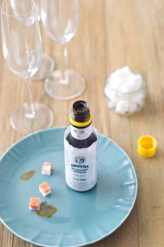 A bottle of bitters with it's cap off sitting on a round blue plate with 3 sugar cubes that have been splashed with bitters. The yellow cap is sitting next to the plate on a wood table with a jar of sugar cubes and some champagne flutes in the background.