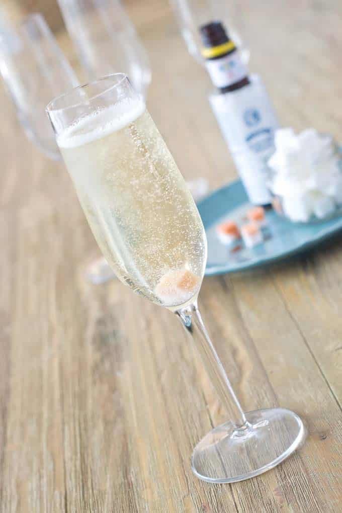 The photo is taken at an angle. There is a champagne flute filled with a bubbly champagne on a wooden table. In the background is a round blue plate with a bottle of bitters on it as well as a jar full of sugar cubes. Also on the place are 3 sugar cubes that have been doused in bitters.
