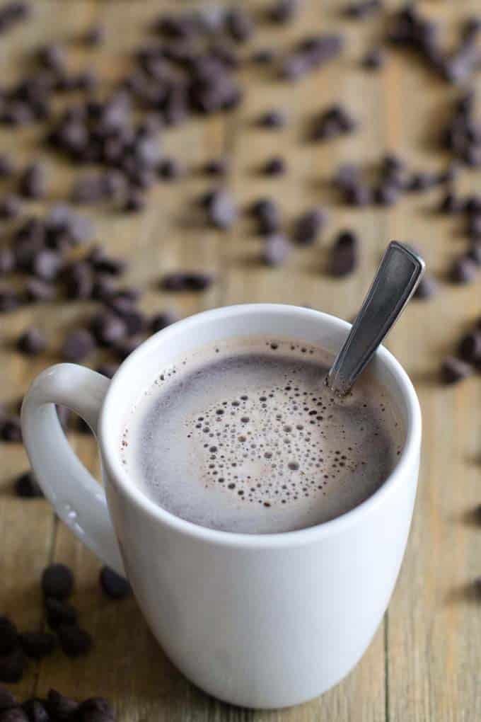 White mug of hot chocolate with a spoon in it. It is on a wooden table that is scattered with chocolate chips.