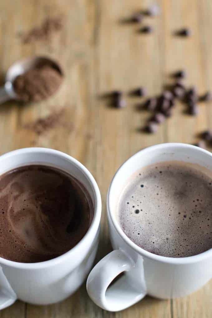 Two white mugs on a wooden table. One mug is filled with hot chocolate and one mug is filled with hot cocoa. In the background are chocolate chips and a spoonful of powdered chocolate.