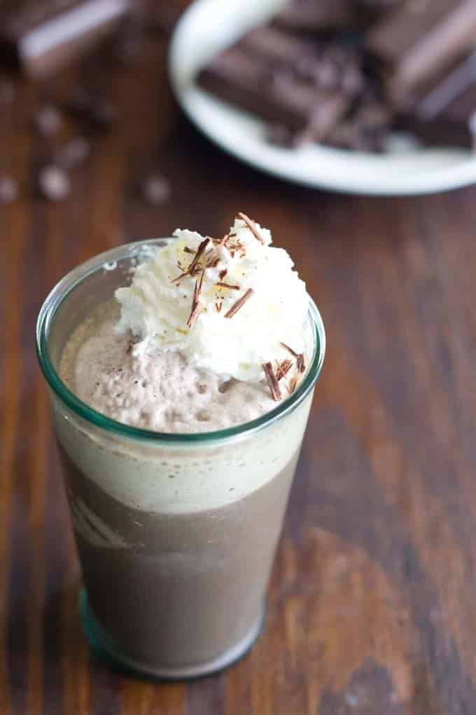 Glass filled with frozen hot chocolate topped with a dollop of whipped cream and sprinkled with shaved chocolate. In the background is a white plate filled with chocolate.