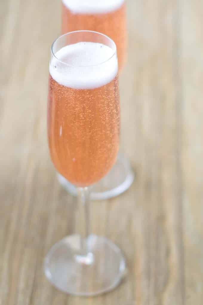 Two champagne flutes filled with bubbling pale pink champagne sitting on a wood table.