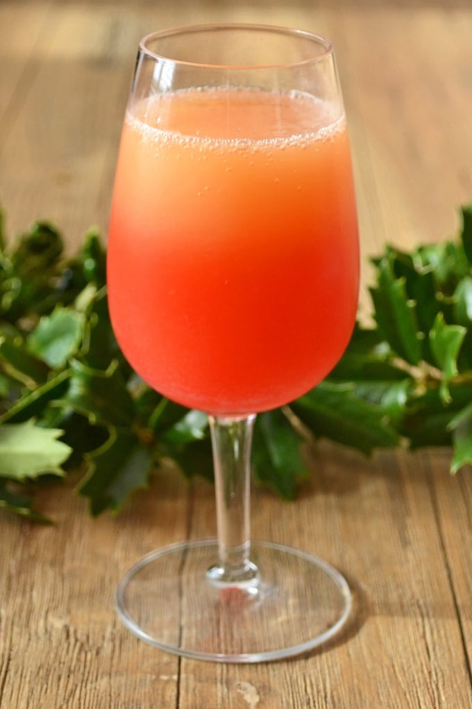 A short stemmed glass filled with a red/orange mimosa. It is sitting on a wood table in front of a garland of holly.