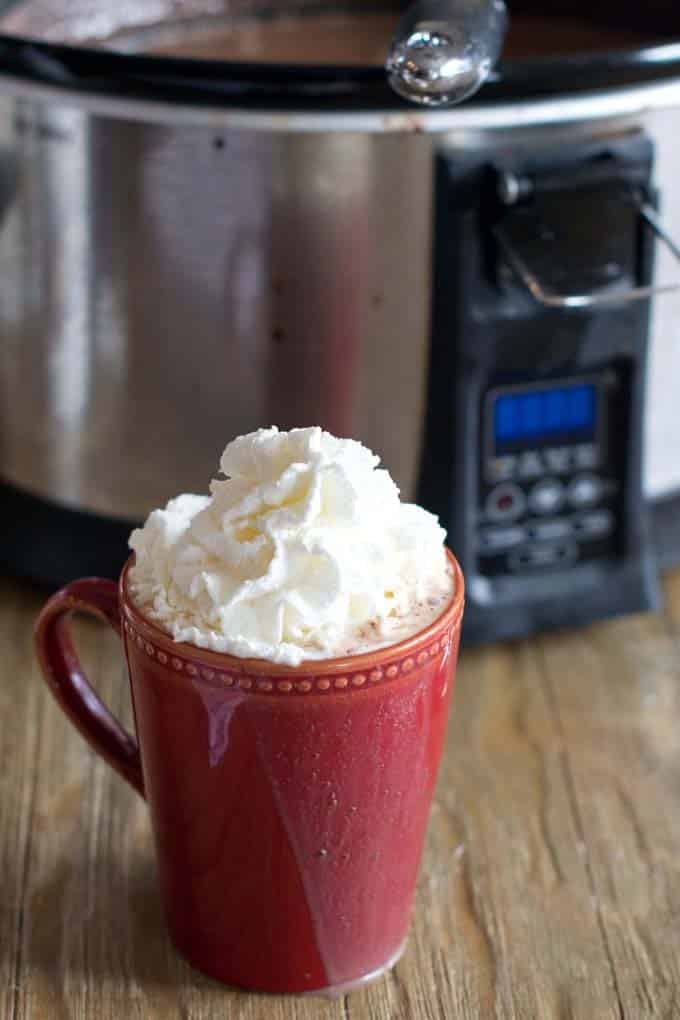 Red mug filled with hot chocolate topped with whipping cream, sitting in front a slow cooking.