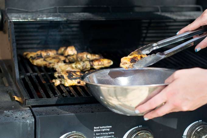 Tongs being used to move chicken from grill to stainless bowl.
