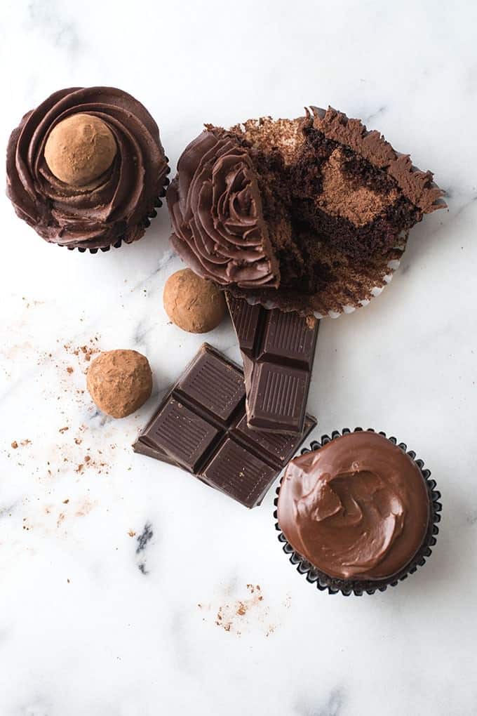 Picture taken from above of three chocolate cupcakes. The first has a chocolate nested in the chocolate frosting, the second is cut in half to reveal a cupcake that is also filled with chocolate the third is a plain chocolate frosted cupcake. Scattered around the cupcakes on the white marble surface is two chocolate truffles and some squares of dark chocolate.