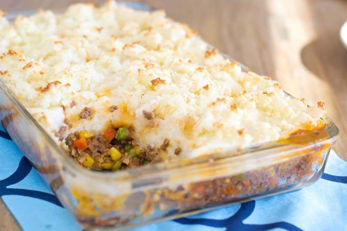 Shepherd's Pie in a rectangular glass pyrex pan with a scoop missing; pan is sitting on a wooden table with a blue table runner.