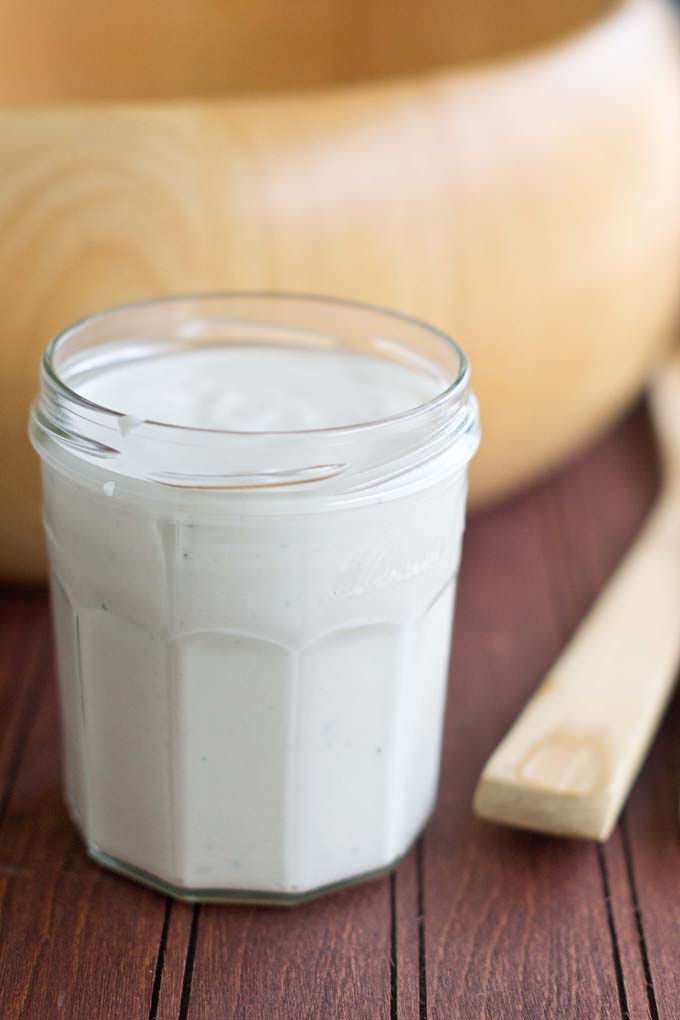 Glass jar filled with white caesar dressing sitting in front of a wooden salad bowl; a salad tosser is sitting next to the glass jar.