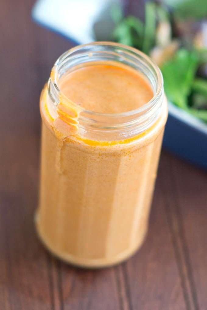 Tall glass jar filled with golden Homemade French Dressing; in the background is a bowl filled with salad greens.