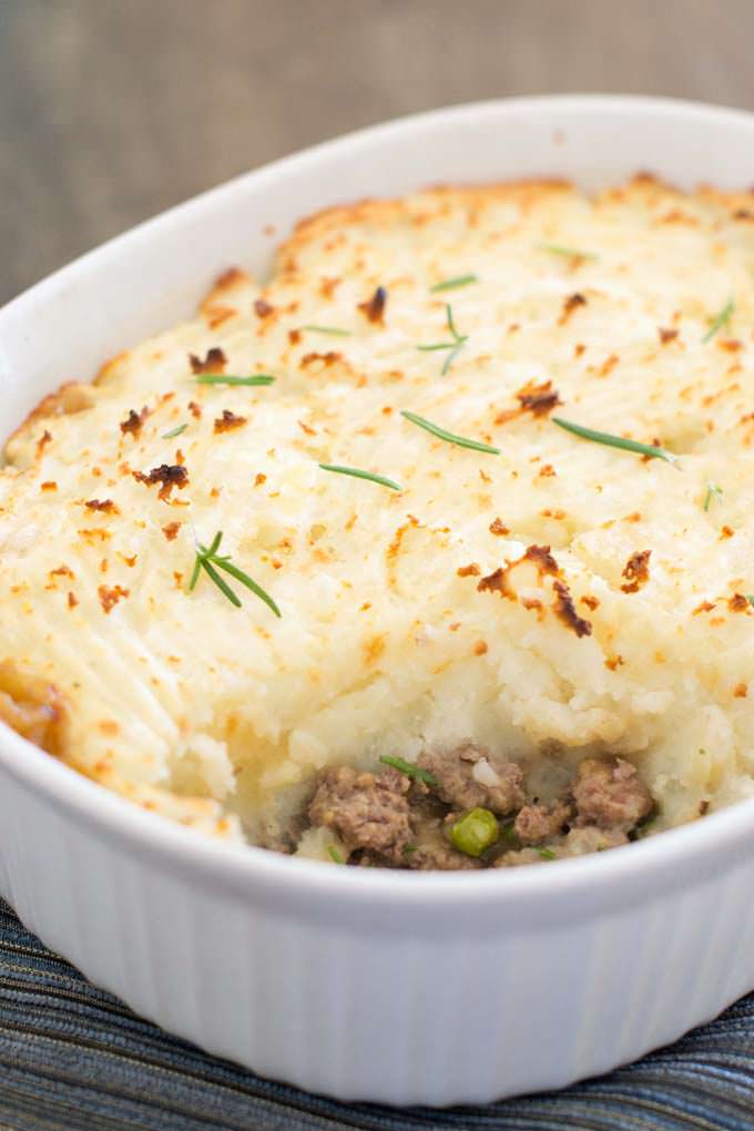 Close up photo of a a white oval casserole dish filled with Shepherd's Pie with a scoop taken out of it so you can see the ground beef and peas underneath the potatoes.