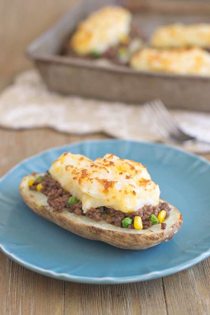 Half of a baked potato sitting on a blue plate on a wooden table filled with Shepard's Pie mix: ground beef, peas, corn and topped with browned mashed potatoes. 