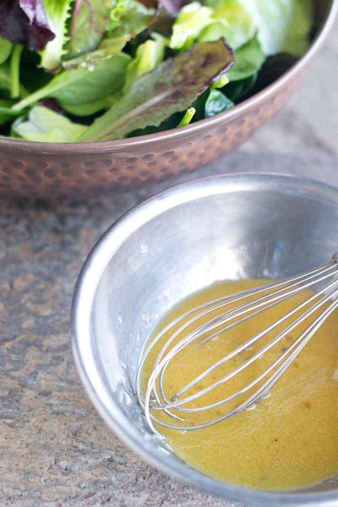 metal mixing bowl with a metal whisk sitting in some vinaigrette; behind it is a brown bowl filled with mixed salad greens.