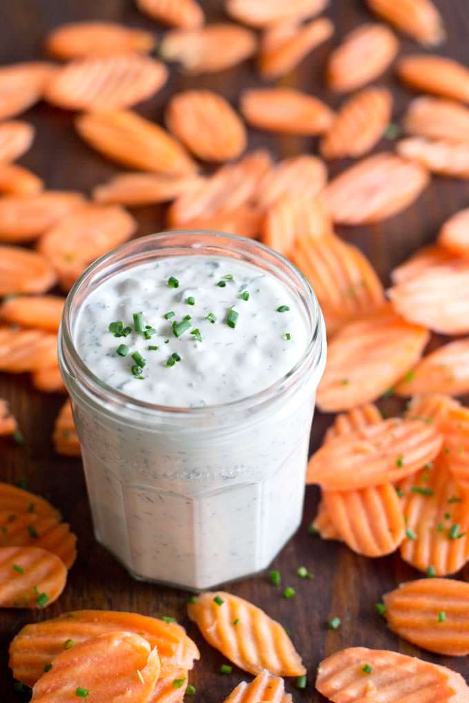 glass jar filled with white creamy ranch dressing with chopped up herbs; garnished with chopped chives; sitting on a wooden surface, surrounded by raw carrot chips that have been sprinkled by chopped chives.