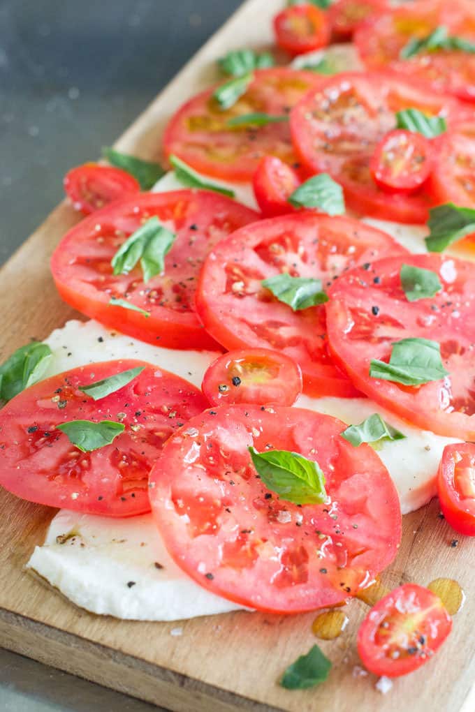 Caprese Salad on a wooden board with slices of tomato, halved cherry tomatoes, slices of fresh mozzarella, and torn basil leaves.