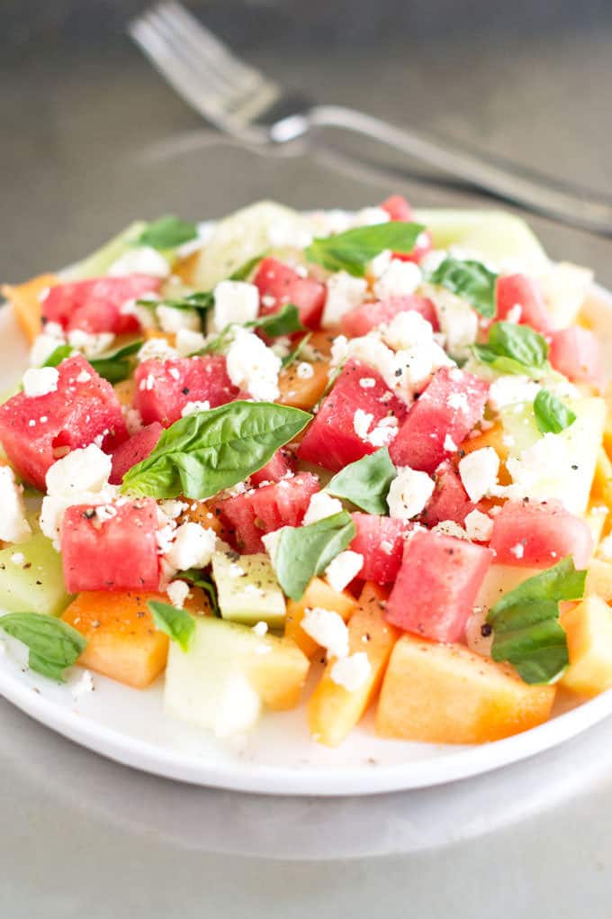 White plate with a salad of various melon chunks, crumbled feta, and fresh basil leaves.