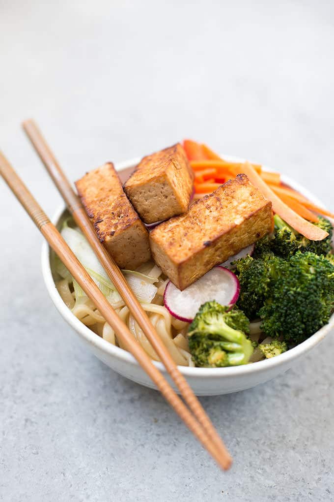 Bowl of asian noodles with broccoli florets, carrot ribbons, green onion slices and marinated cooked tofu chunks on top of the noodles. Chop sticks are resting across the top of the bowl.