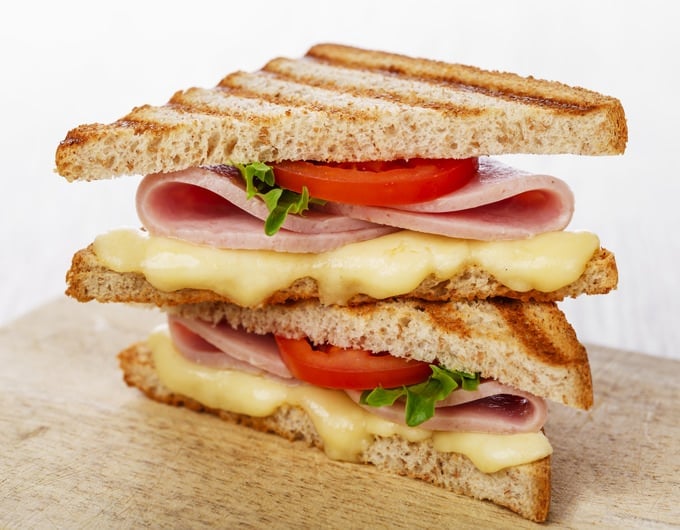 Half of a triple decker sandwich on a wooden table with a white background. The sandwich contains melted cheese, tomato, lettuce and folded ham slices.