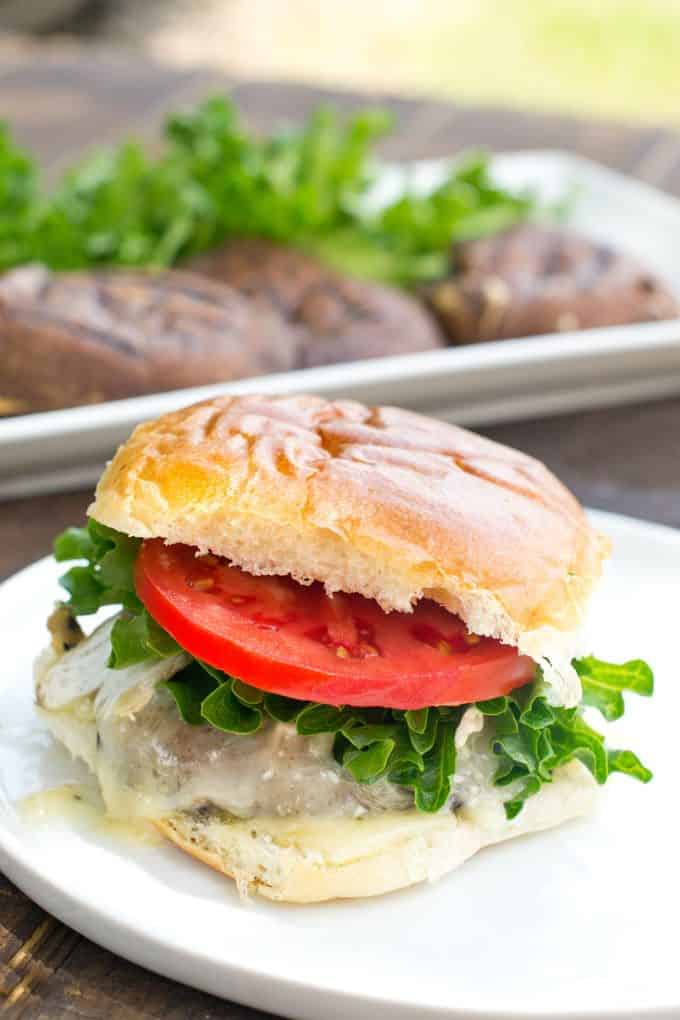 Grilled portobello mushroom burger with melted cheese, lettuce and tomato on a white bun; in the background is a rectangular white plate filled with Grilled Portobello Mushrooms.