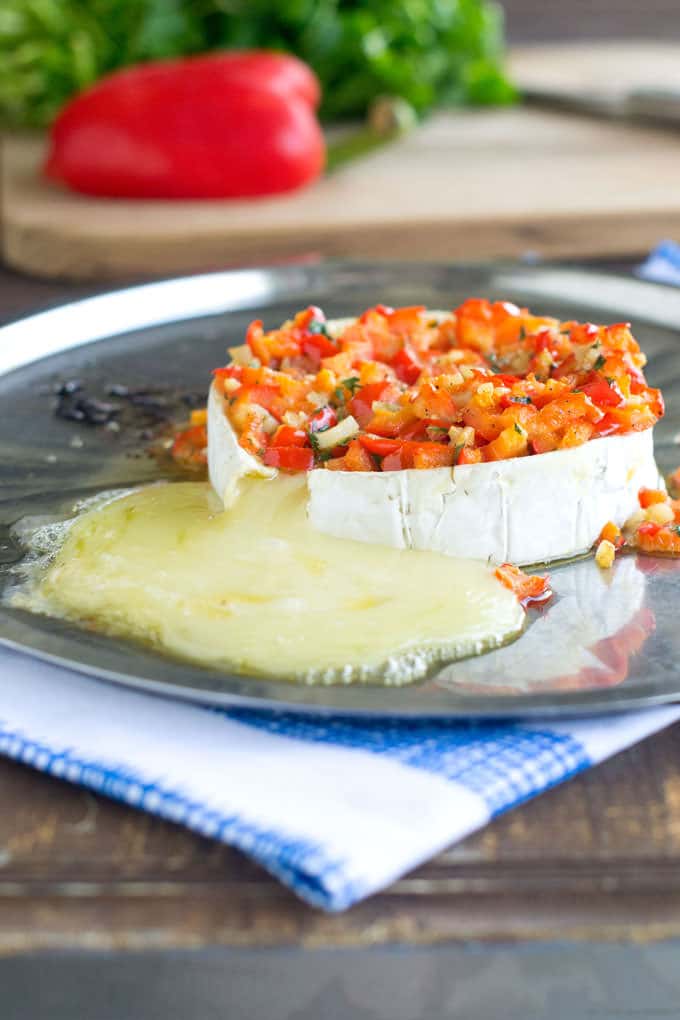 Wheel of brie cheese topped with diced red peppers cooked with garlic and parsley. The brie has been baked and she soft cheese is oozing out onto the silver platter it is on.