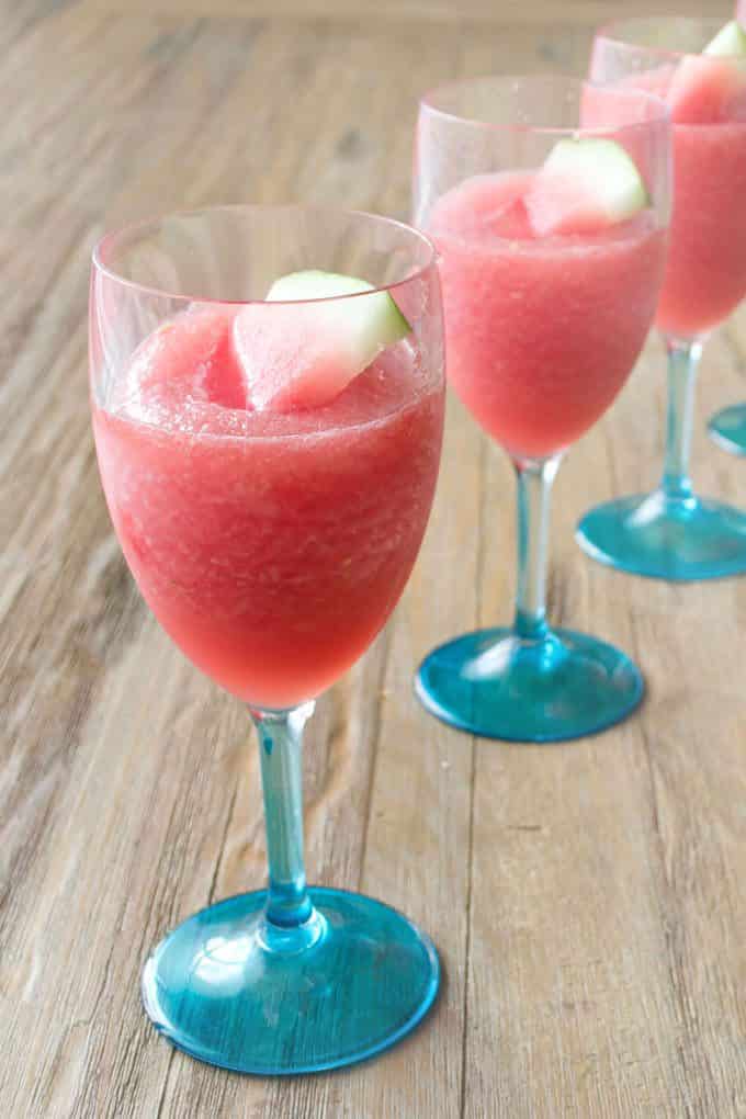 A line of three wine glasses with blue stems filled with Watermelon Lemonade Wine Slush that has a small wedge of watermelon with the rinds in it.