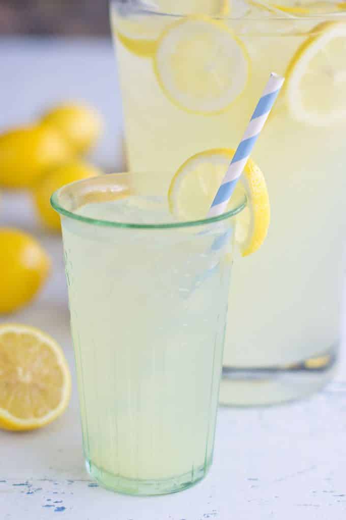 tall glass filled with lemonade and a blue and white stripped straw. There is a slice of lemon on the edge of the glass. In the background is a pitcher of lemonade and some whole lemons and one half of a lemon.