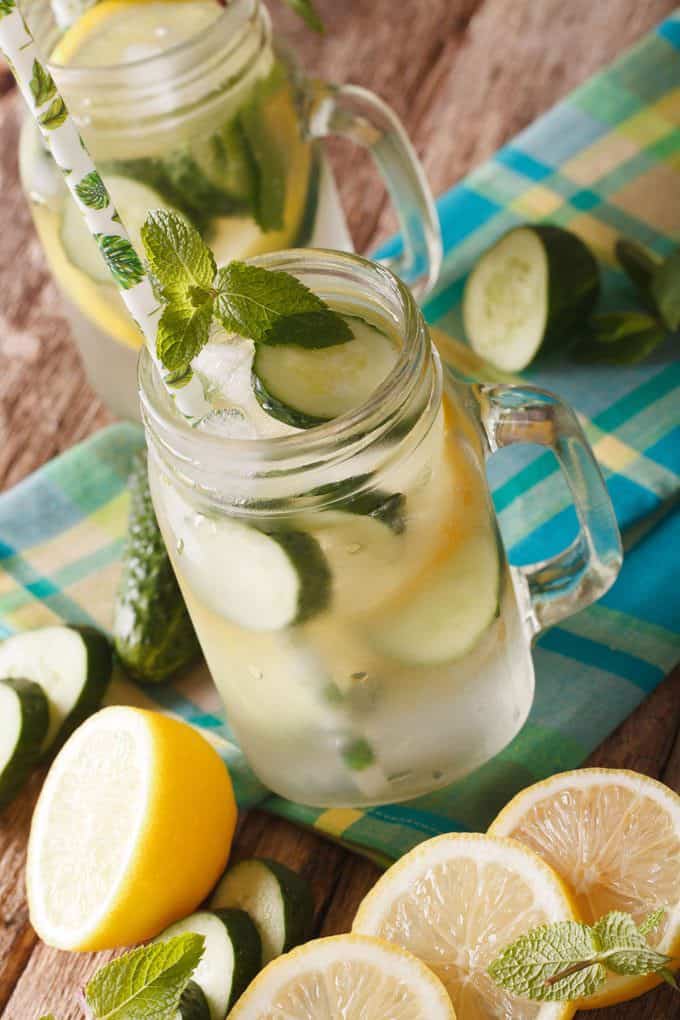 Mason jar mug filled with lemonade that has slices of cucumber, lemons, a straw and a sprig of mint in it. It is sitting on a blue, green and yellow plaid napkin with slices of cucumbers and slices of lemons scattered around it.