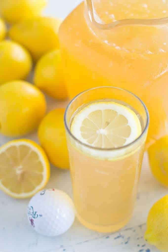 Tall glass of dark looking lemonade with a slice of lemon floating on the top surrounded by lots of whole lemons, one half of a lemon and a golf ball.