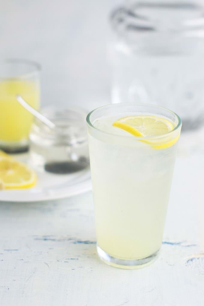 Tall glass of lemonade with a slice of lemon floating in it. In the background is a round white plate with a jar of simple syrup with a spoon sticking out of it, an slice of lemon and tall glass jar of lemon juice.