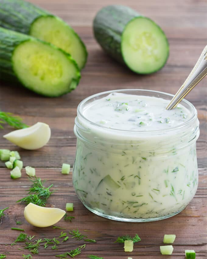 Tzatziki sauce in a short jar with garlic cloves, chopped cucumber, and dill scattered around, cucumbers in the background. The words Authentic Tzatziki Sauce appear on the image.