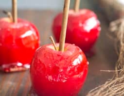 Three red apples coated in candy with rustic sticks coming out of the tops. There's grassy hay beside them.